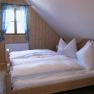 Zimmer, © Ferienhaus Hintereck Gemütliches Schlafzimmer mit Holzwänden, einem Doppelbett und einem Fenster mit blau karierten Vorhängen.