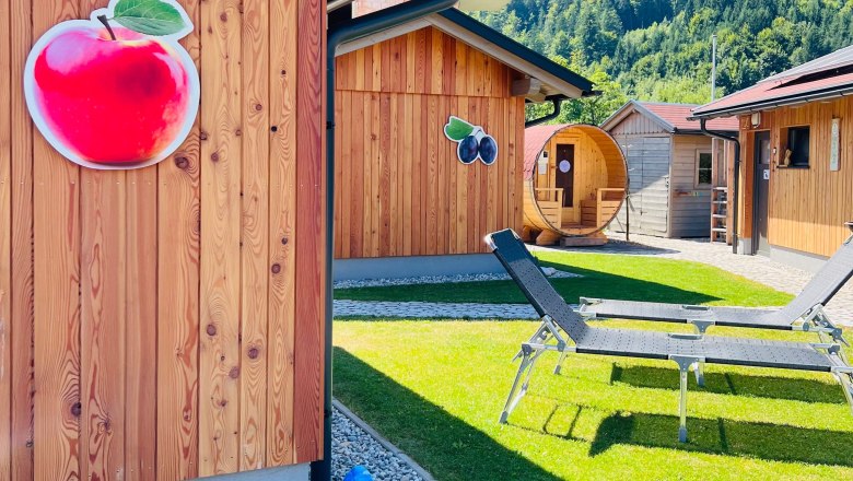 Meine kleine Auszeit -Die Schlafhütten, © Melanie Heim Holzhütten mit Obstdekoration und Liegestühlen auf einer Wiese, umgeben von Bergen.