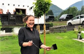 Feiern im Erlauftalerhof, © Erlauftalerhof Eine Frau steht lachend an einer Feuerstelle im Freien, umgeben von grüner Landschaft und einem Gebäude im Hintergrund.