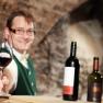 Gasthof Teufl, © weinfranz.at Ein Mann in traditioneller Kleidung bietet ein Glas Rotwein an, im Hintergrund zwei Weinflaschen.