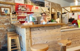 Edelweißlounge, © Franz Heher Eine Frau steht hinter einer rustikalen Holztheke in einer gemütlichen Lounge mit Holzhockern und Steinwand.