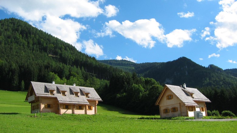 Feriendorf Lassing-Hochkar, © Friedrich Tippelreither Zwei Holzhäuser auf einer grünen Wiese vor bewaldeten Bergen und blauem Himmel.