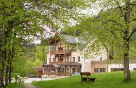 Fastenhaus Maria Seesal, © Fastenhaus Ein großes Gebäude mit Holzbalkonen, umgeben von grünen Bäumen und einer Wiese mit einer Bank im Vordergrund.