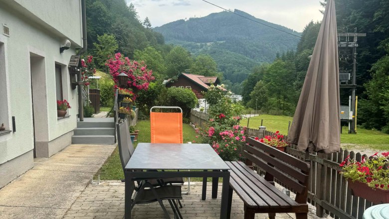 Ferienwohnung Schindlegger, © Familie Schindlegger Terrasse einer Ferienwohnung mit Tisch, Stühlen und Blumen, umgeben von grünen Hügeln und Wald.