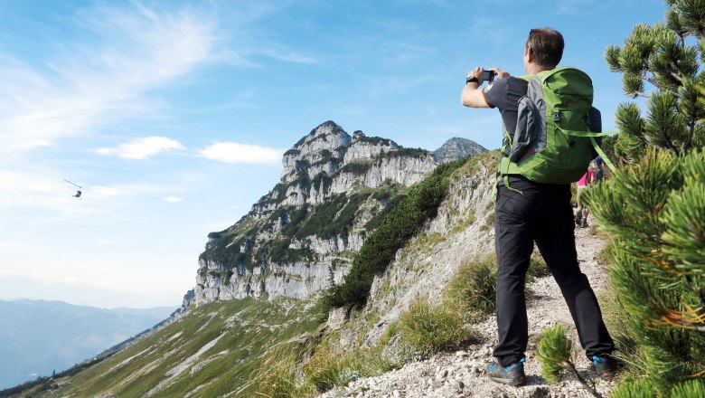Ötscher - Rauher Kamm, © weinfranz.at Ein Wanderer mit grünem Rucksack fotografiert eine Berglandschaft mit einem Hubschrauber am Himmel.