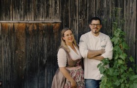 Wirtsleute Sabrina und Markus Stadler, © Niederösterreich Werbung/Julius Hirtzberger Ein Paar steht vor einer Holzwand, sie trägt ein Dirndl, er eine Kochjacke.