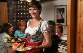 Wanderrast Hochschlag, © zVg Eine Frau in traditioneller Kleidung serviert Essen in einem gemütlichen Raum.