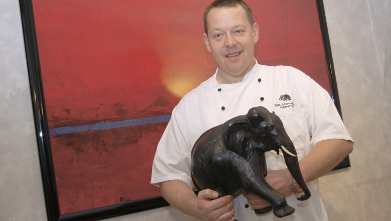 Restaurant zum schwarzen Elefanten Heinz Eisbacher, © diewerbetrommel.at Ein Koch in weißer Uniform hält eine schwarze Elefantenfigur vor einem roten Gemälde.