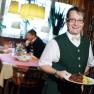 Gasthof Teufl, © Gasthof Teufl Ein Kellner in grüner Weste serviert ein Gericht in einem Restaurant. Im Hintergrund sitzen zwei Männer an einem Tisch.