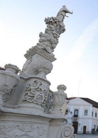 Johannes Nepomuk Säule, © schwarz-koenig.at Nahaufnahme einer kunstvoll verzierten Säule mit Skulpturen und Ornamenten, im Hintergrund Gebäude und blauer Himmel.