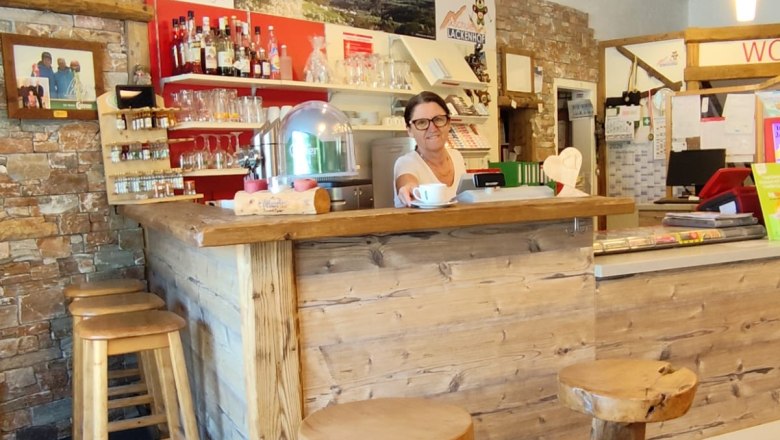 Edelweißlounge, © Franz Heher Eine Frau steht hinter einer rustikalen Holztheke in einer gemütlichen Lounge mit Holzhockern und Steinwand.