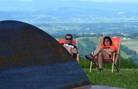 Rund um Schau, © Kulturpark Eisenstraße Zwei Personen entspannen in Liegestühlen auf einer Wiese mit Blick auf eine weite Landschaft.