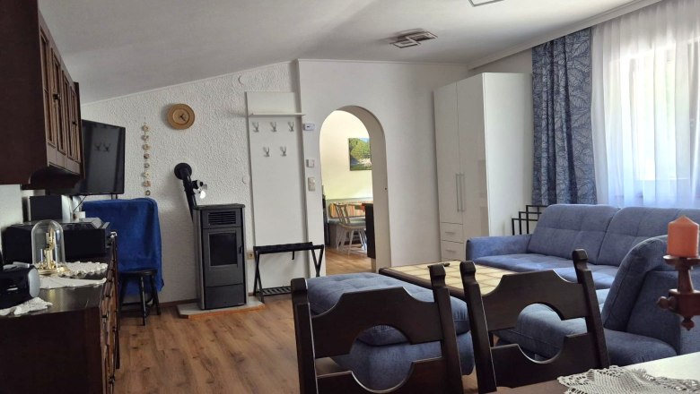 Ferienwohnung Schindlegger, © Familie Schindlegger Gemütliches Wohnzimmer einer Ferienwohnung mit blauen Sofas, Holzmöbeln und einem Kamin. Ein Esstisch ist im Hintergrund sichtbar.