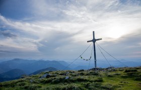 Ein majest&auml;tisches Gipfelkreuz thront &uuml;ber den sanften H&uuml;geln, umgeben von einer atemberaubenden Berglandschaft. Die Wolken ziehen sanft &uuml;ber den Himmel und verleihen der Szenerie eine friedliche Atmosph&auml;re, w&auml;hrend die frische Bergluft zum Wandern einl&auml;dt.