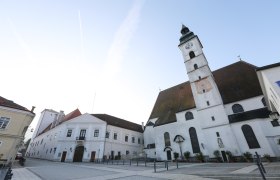 Pfarrkirche mit Uhrturm und angrenzenden Gebäuden auf einem leeren Platz.