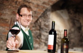 Ein Mann in traditioneller Kleidung bietet ein Glas Rotwein an, im Hintergrund zwei Weinflaschen.