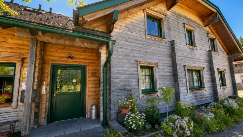 Ein traditionelles Holzhaus mit gr&uuml;nen Fensterrahmen und einer gr&uuml;nen T&uuml;r, umgeben von Blumen und Pflanzen, unter einem klaren blauen Himmel.