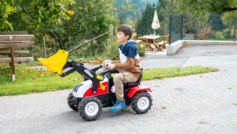 Ein Kind f&auml;hrt auf einem kleinen Trettraktor mit Schaufel auf einem Weg im Freien.