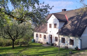 Ein traditionelles Landhaus mit wei&szlig;en W&auml;nden und braunen Fenstern, umgeben von gr&uuml;nen B&auml;umen und einem gepflegten Garten mit einem Picknicktisch.