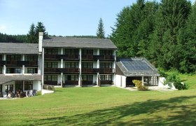 Hotelgeb&auml;ude mit Solarpanelen und gr&uuml;ner Wiese im Vordergrund, umgeben von Wald.