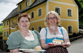 Zwei Frauen in traditioneller Kleidung halten Teller mit Speisen vor einem gelben Geb&auml;ude.