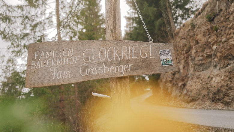 Holzschild mit der Aufschrift 'Familienbauernhof Glockriegl Fam. Grasberger' an einem Waldweg.
