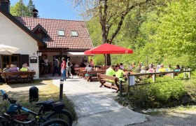 Eine Bergh&uuml;tte mit Terrasse, auf der Menschen an Tischen sitzen. Fahrr&auml;der sind im Vordergrund zu sehen, umgeben von gr&uuml;ner Natur.