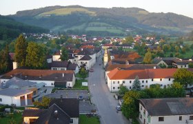 Luftaufnahme eines Dorfes mit roten Dächern und grüner Landschaft im Hintergrund.