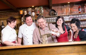 Eine Gruppe von Menschen hinter einer Bar, die gemeinsam lachen und eine Flasche in einen Krug gießen.