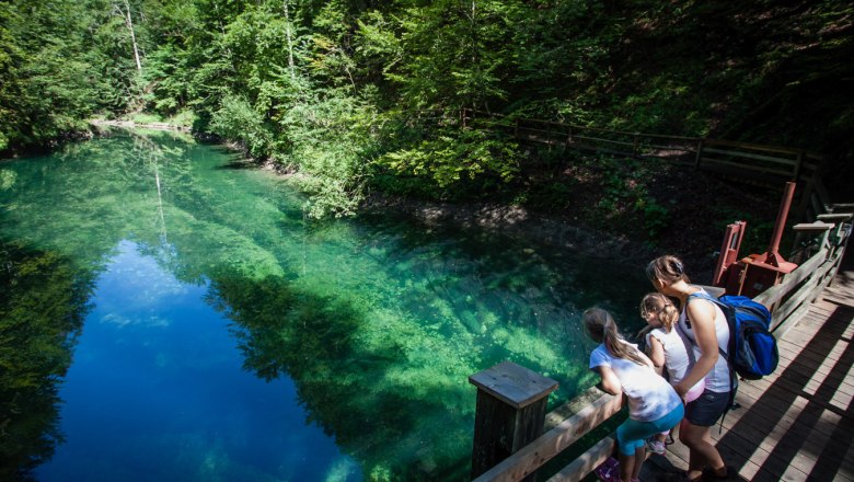 Eine Frau und zwei Kinder schauen von einem Holzsteg auf einen klaren, grünen Fluss in einem Wald.