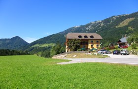 Ein gelbes Gebäude mit Balkon und Blumen, umgeben von grünen Wiesen und Bergen unter blauem Himmel.