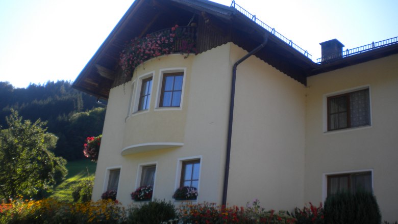 Gelbes Haus mit Blumen und Balkon vor bewaldetem Hügel.