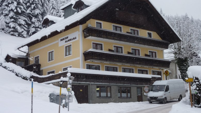Ein verschneites Gebäude mit der Aufschrift 'Pension Panorama' in einer winterlichen Landschaft.