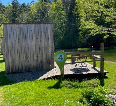 Eine kleine Holzh&uuml;tte mit Terrasse und Tisch im Gr&uuml;nen, umgeben von B&auml;umen, direkt am Teich gelegen.