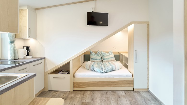 Kompaktes Apartment mit integriertem Bett unter einer Dachschr&auml;ge, K&uuml;chenzeile und Fernseher.