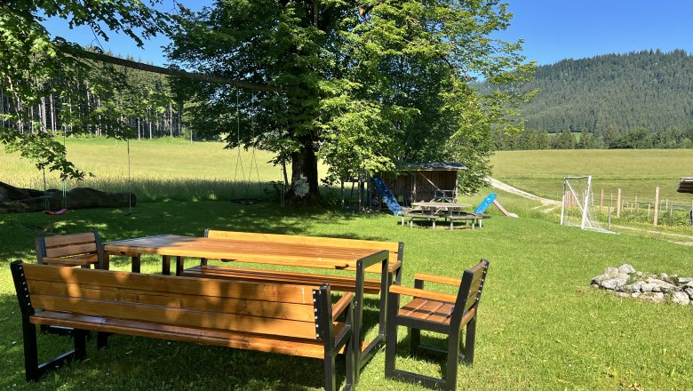 Ein Garten mit Holztisch und B&auml;nken, umgeben von B&auml;umen und Wiesen, mit einem Spielplatz im Hintergrund.