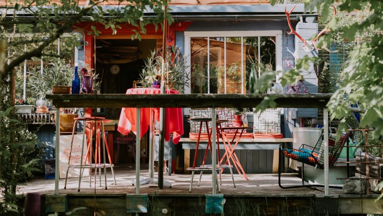Bunte Terrasse vor der Location mit roten St&uuml;hlen und Tisch, umgeben von Pflanzen und Dekorationen.