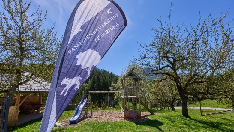 Spielplatz mit Rutsche und Schaukel im Garten eines Bauernhofs, umgeben von B&auml;umen und Wiesen.