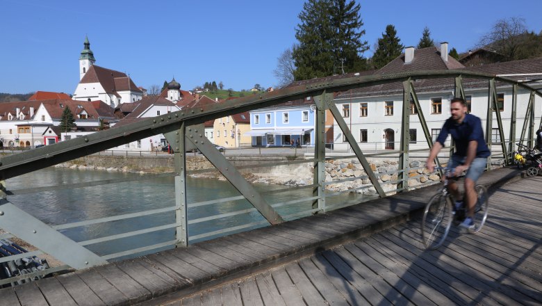 Ein Radfahrer &uuml;berquert die T&ouml;pperbr&uuml;cke in einer malerischen Stadt mit Kirche und bunten H&auml;usern im Hintergrund.