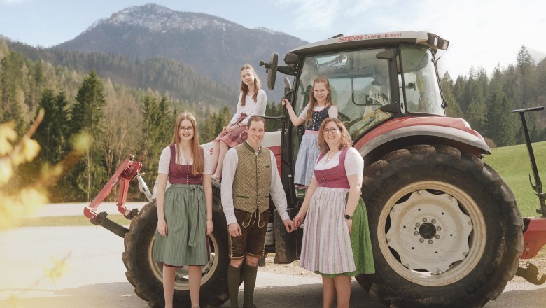 Eine Familie in traditioneller Tracht steht vor einem Traktor in einer l&auml;ndlichen Berglandschaft.