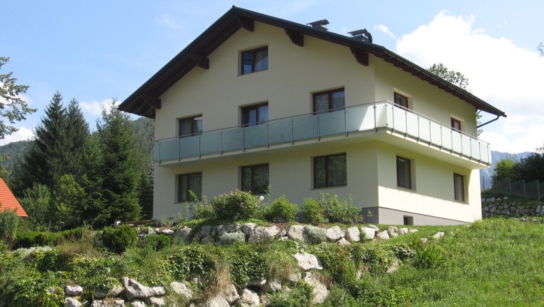Ein zweistöckiges Haus mit Balkon in einer grünen, hügeligen Landschaft.