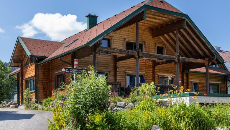 Ein traditionelles Holzhaus mit einem roten Dach, umgeben von bunten Blumen und gr&uuml;nen Pflanzen, unter einem blauen Himmel.