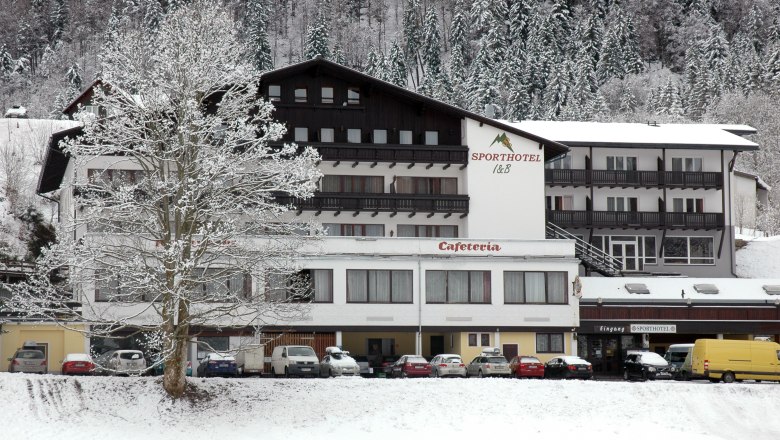 Ein verschneites Hotelgebäude mit der Aufschrift 'Sporthotel' und 'Cafeteria', umgeben von schneebedeckten Bäumen und geparkten Autos.
