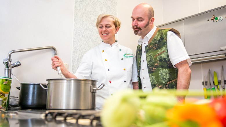 Zwei Personen in einer K&uuml;che, eine r&uuml;hrt in einem Topf, die andere schaut zu.