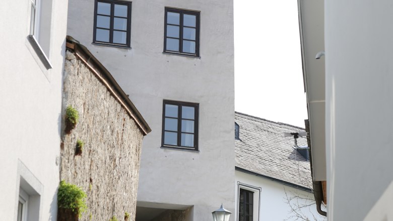 Zwei Personen gehen eine gepflasterte Gasse entlang, vorbei an einem historischen Turm mit mehreren Fenstern.