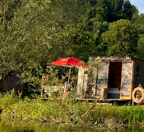 Schlafbox mit rotem Sonnenschirm am Ufer des Neubrucker Teichs, umgeben von B&auml;umen.