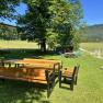 Ein Garten mit Holztisch und B&auml;nken, umgeben von B&auml;umen und Wiesen, mit einem Spielplatz im Hintergrund.