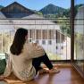 Eine Frau sitzt auf einer Fensterbank und blickt auf eine l&auml;ndliche Landschaft mit Bergen im Hintergrund.