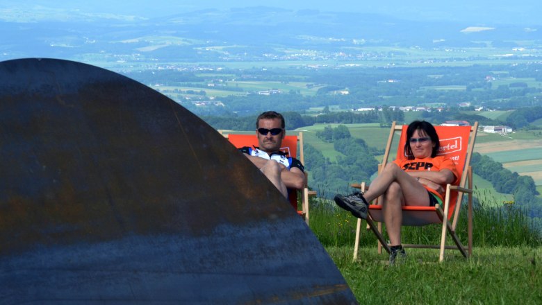 Zwei Personen entspannen in Liegestühlen auf einer Wiese mit Blick auf eine weite Landschaft.
