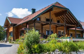 Ein traditionelles Holzhaus mit einem roten Dach, umgeben von bunten Blumen und gr&uuml;nen Pflanzen, unter einem blauen Himmel.